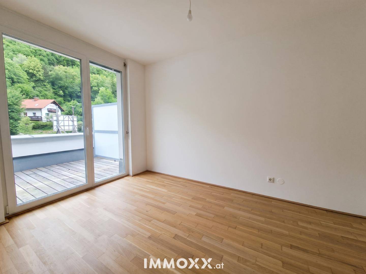 Unmöblierter Raum mit Holzboden und breiter Glasfront, die auf einen Balkon führt und Bergblick bietet.
