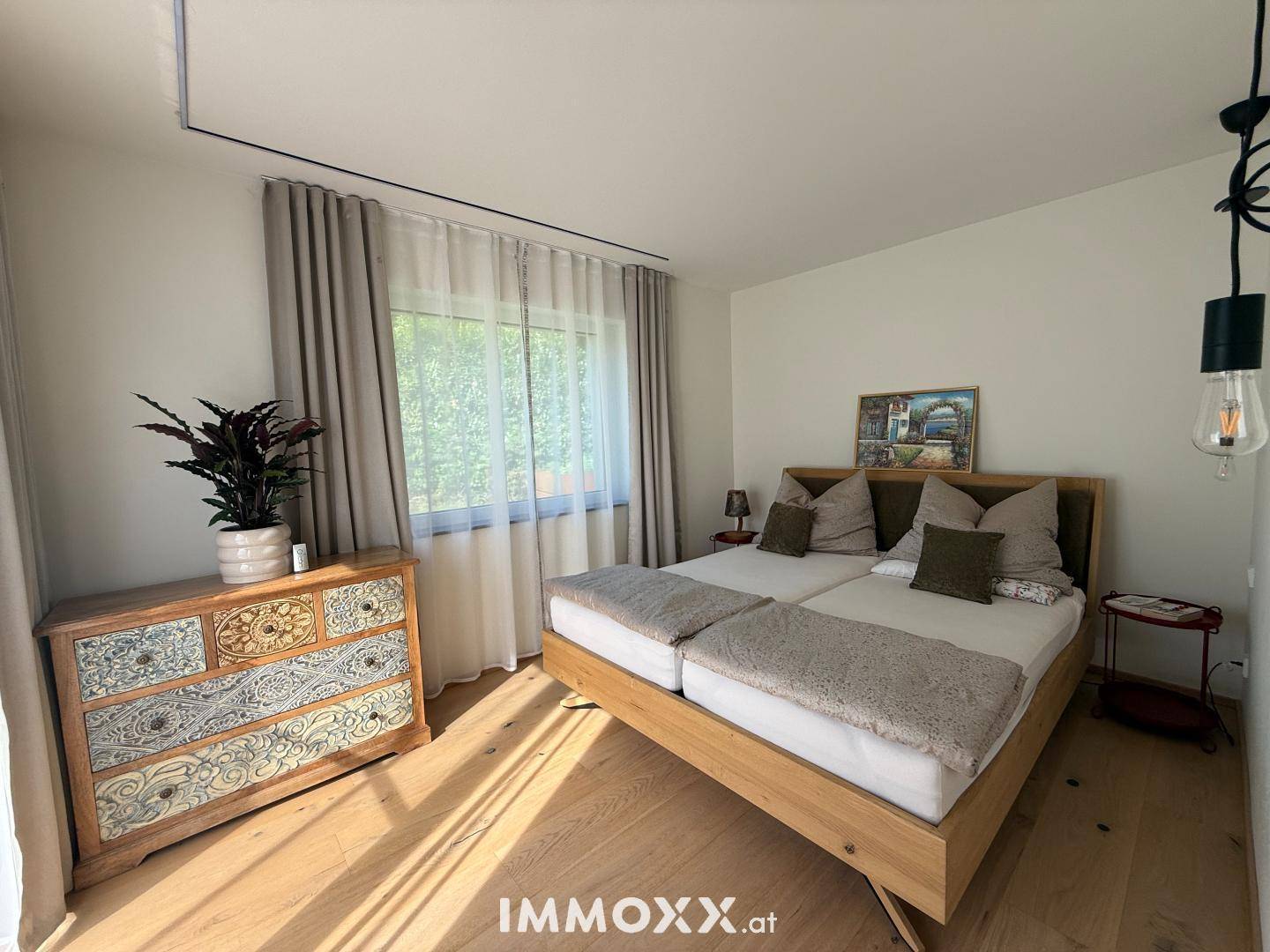 Helles Schlafzimmer mit Holzbett, dekorativer Kommode und Fenster mit Blick ins Grüne.