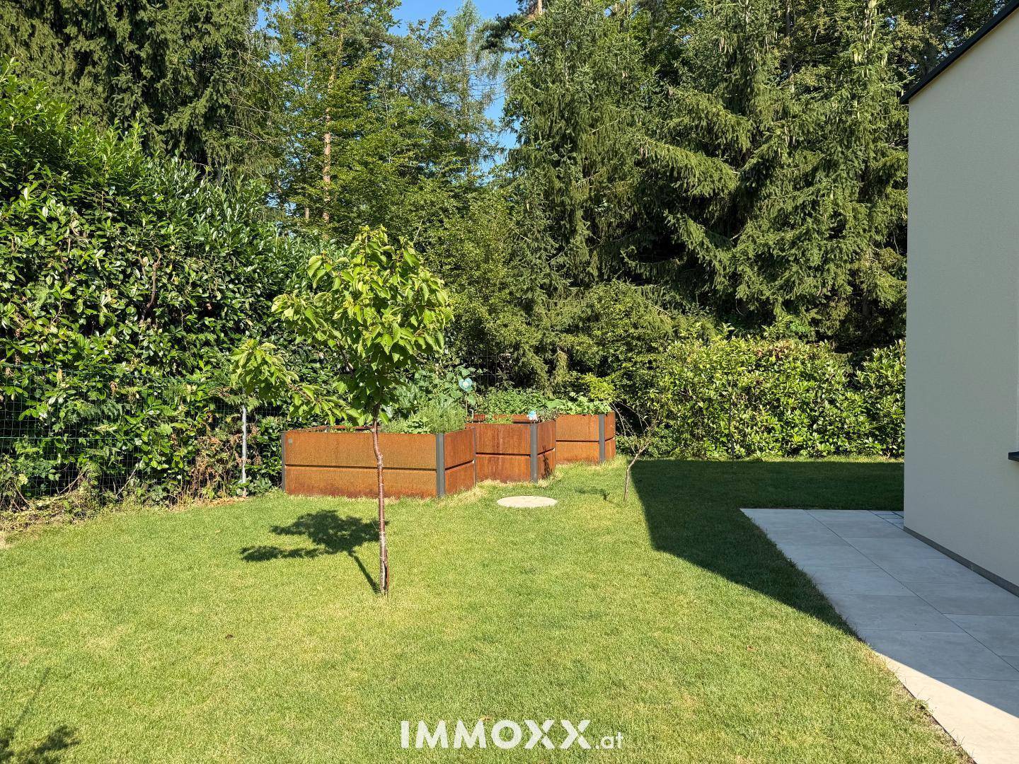 Gepflegter Garten mit Rasenfläche, Hochbeeten und üppiger Bepflanzung im Hintergrund.
