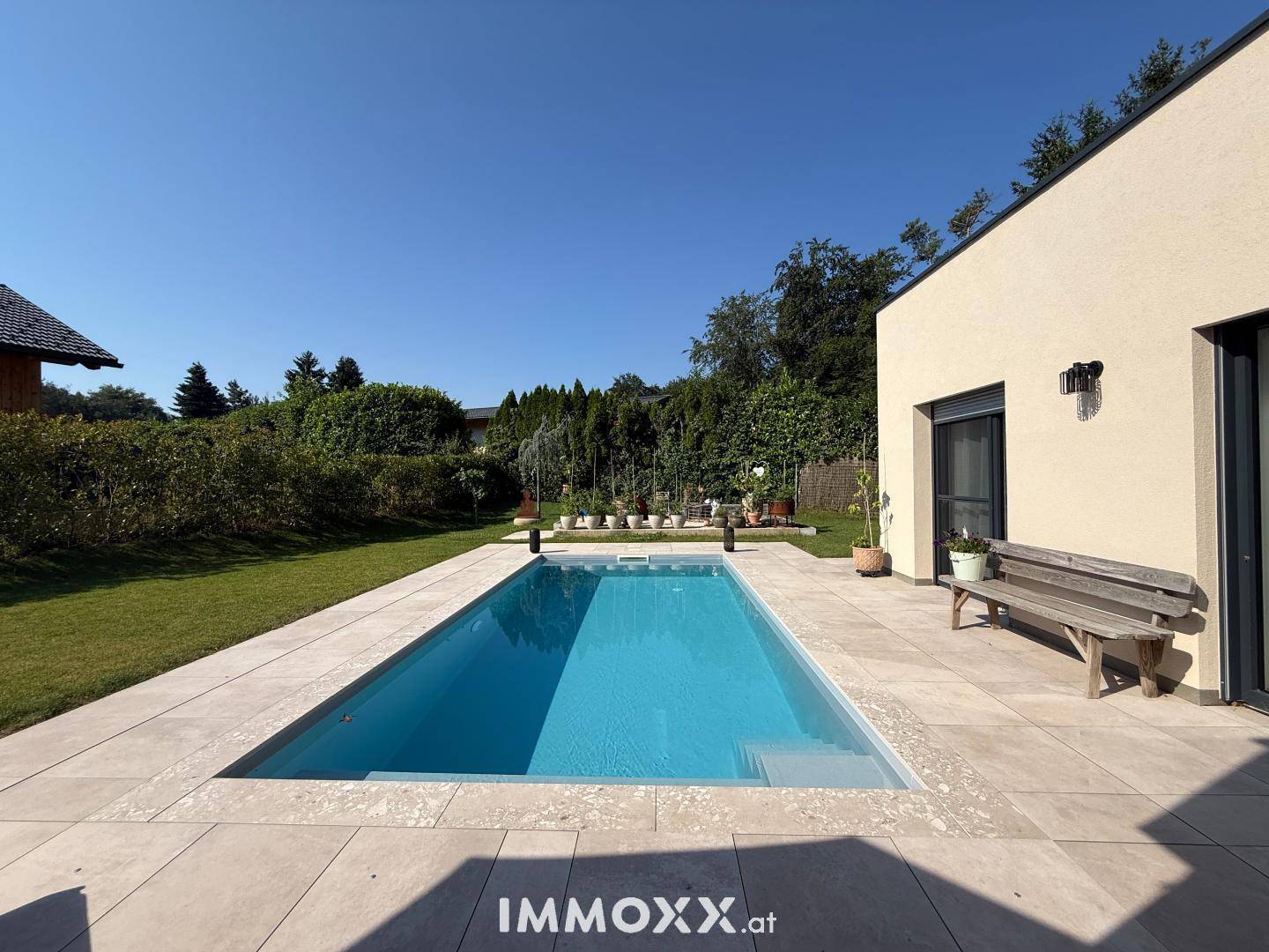 Langer Swimmingpool mit gefliestem Randbereich und angrenzendem grünen Garten unter blauem Himmel.