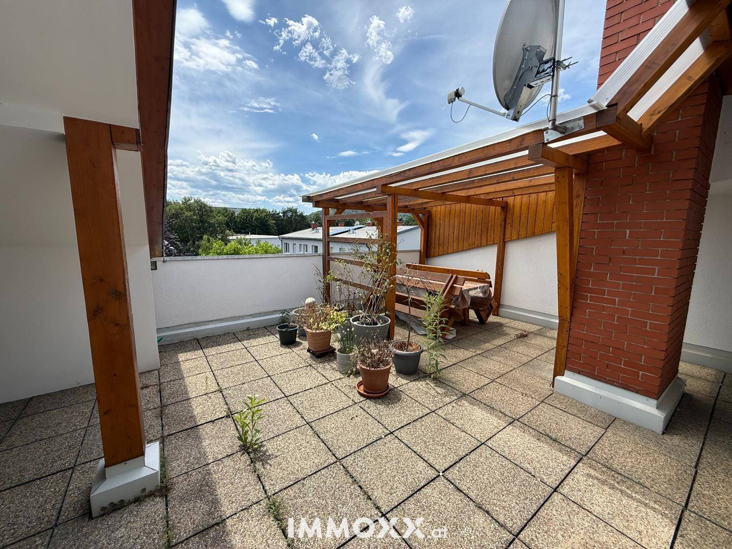 Dachterrasse mit Fliesenboden, Holzpergola und einigen Pflanzen unter blauem Himmel.