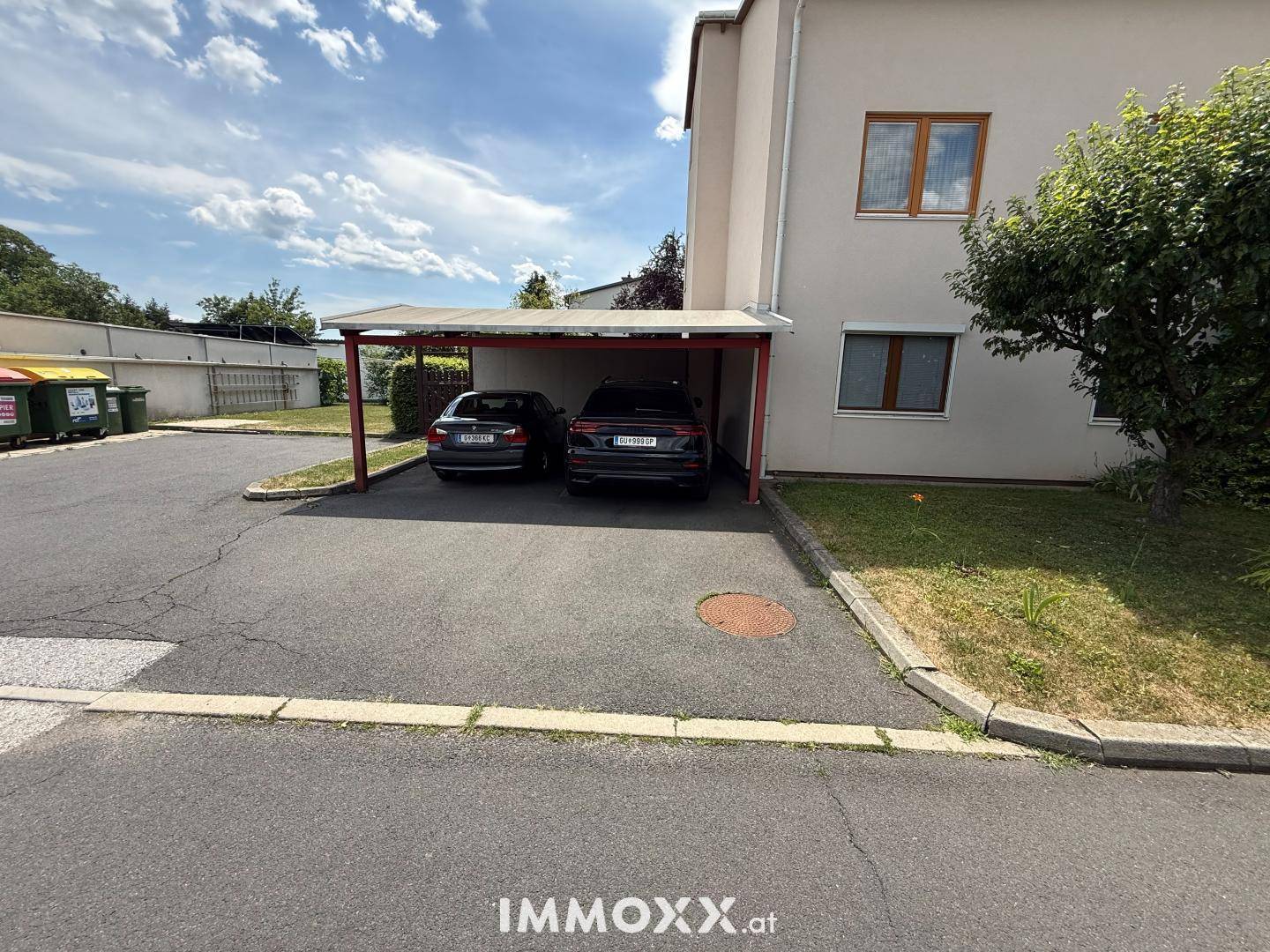 Überdachter Carport mit zwei Fahrzeugen neben einem Wohngebäude und asphaltierter Zufahrt.