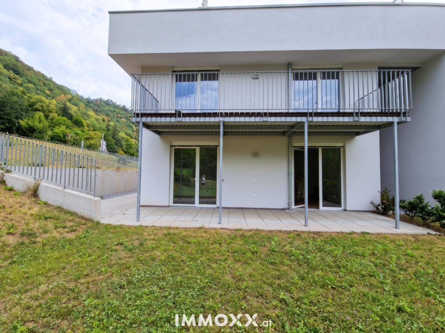Rückseite des modernen Gebäudes mit Balkon, Terrasse und angrenzender Rasenfläche vor bewaldetem Hügel.