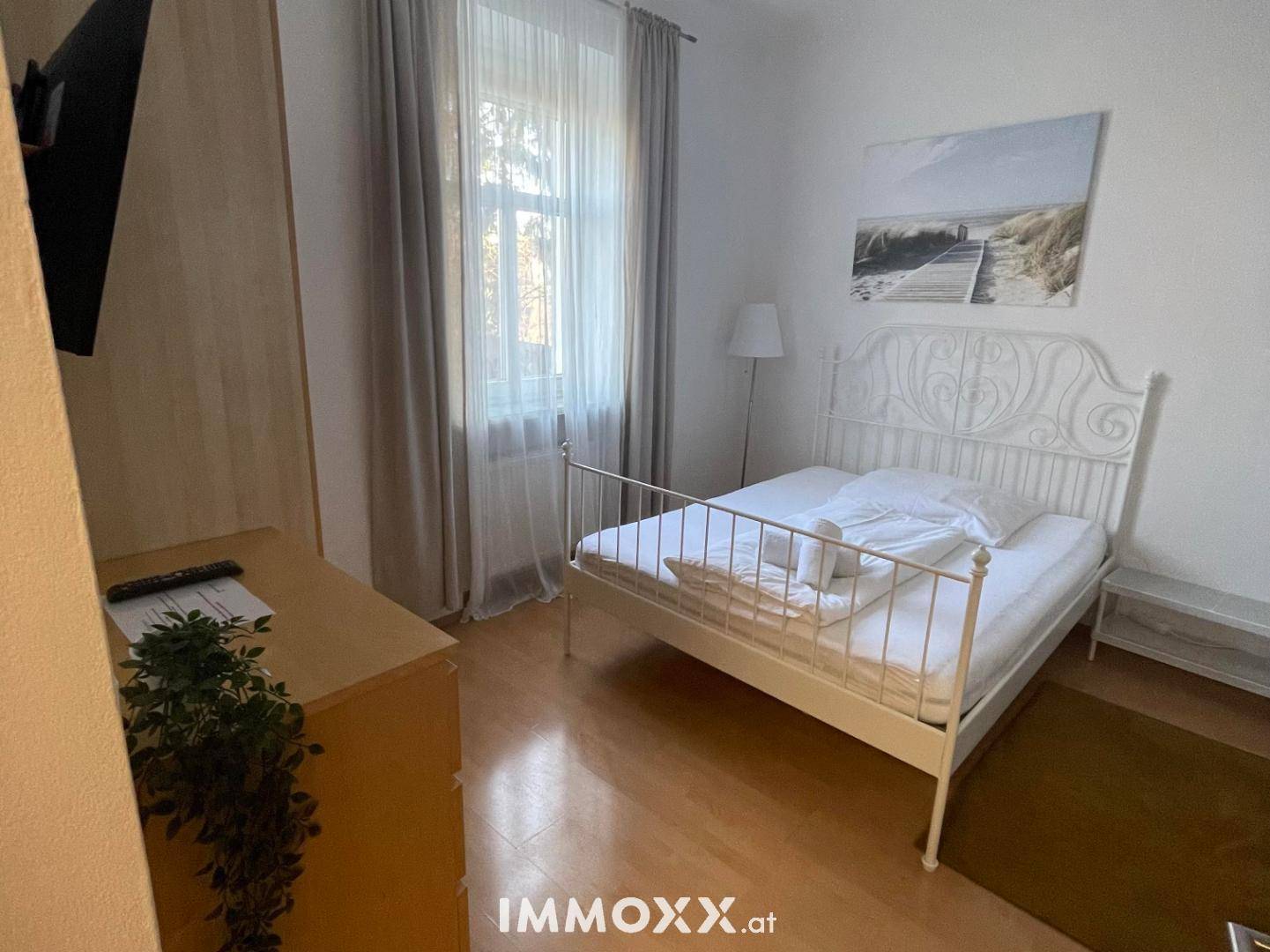 Gemütliches Schlafzimmer mit weißem Metallbett, Holzkommode und Fenster mit hellen Vorhängen.