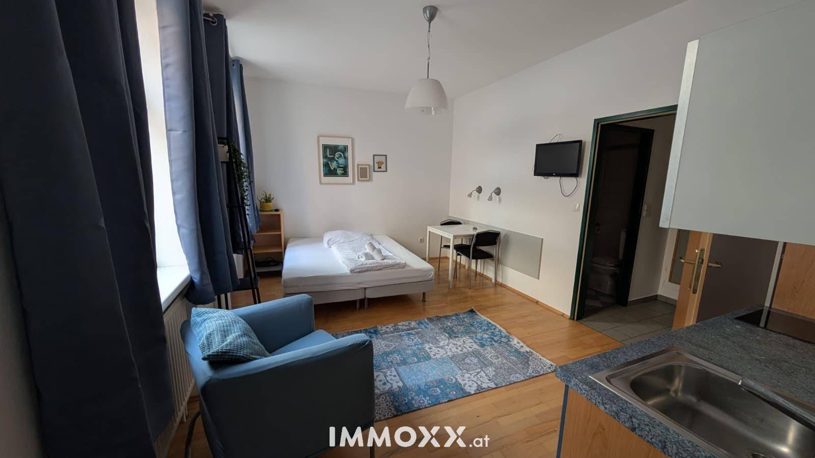 Wohn- und Schlafbereich mit Bett, blauem Sessel und integrierter Kochnische, modern gestaltet.