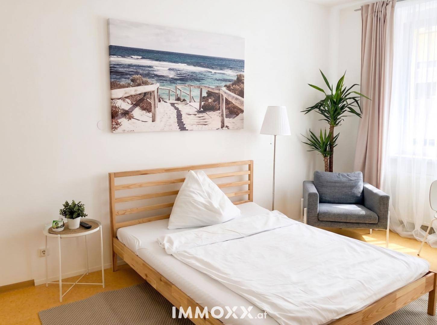 Helles Schlafzimmer mit Holzbett, bequemen Sessel und dekorativem Wandbild, gut beleuchtet.