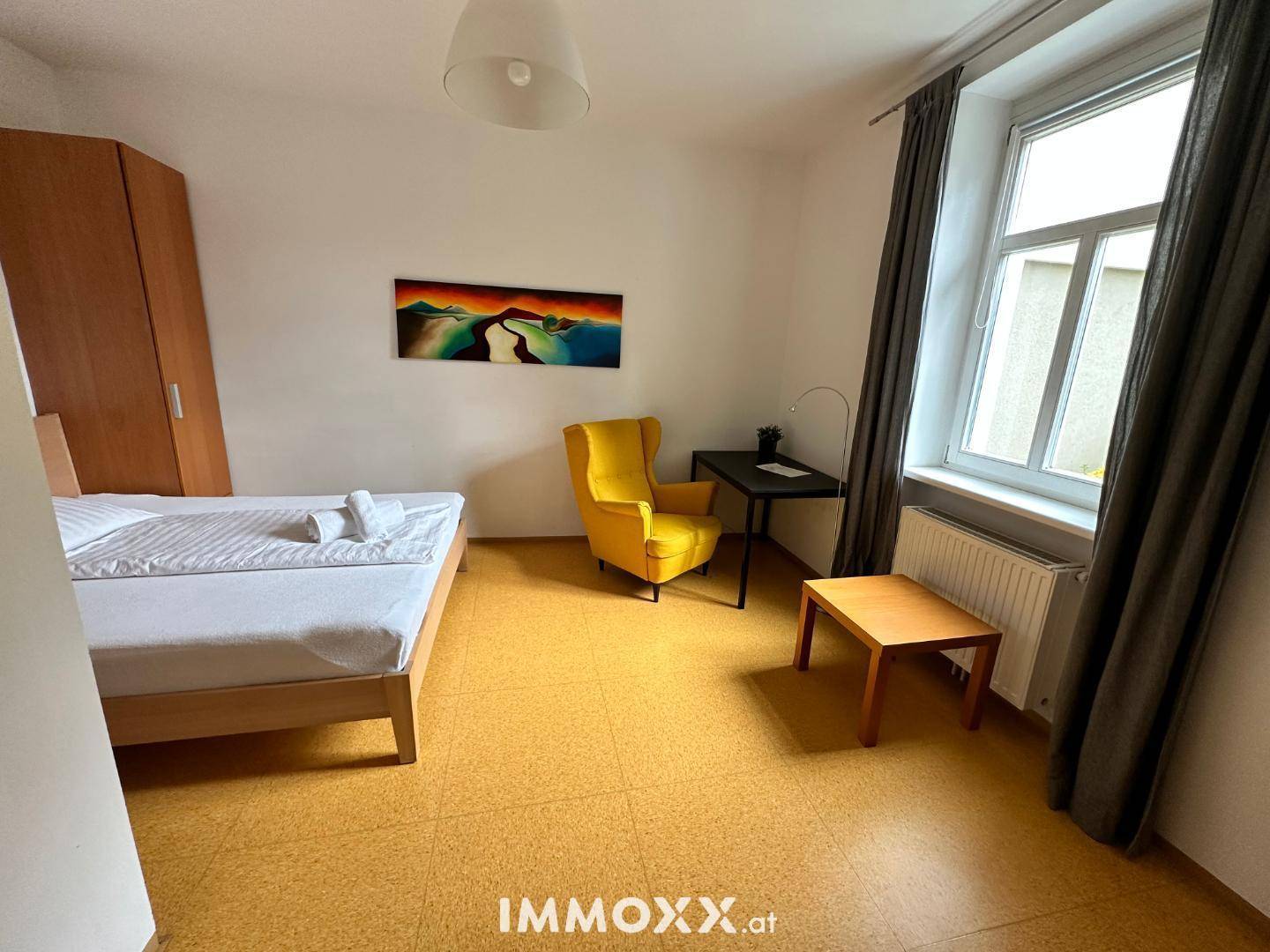 Schlafzimmer mit Holzbett, auffälligem gelben Sessel und kleinem Schreibtisch am Fenster.