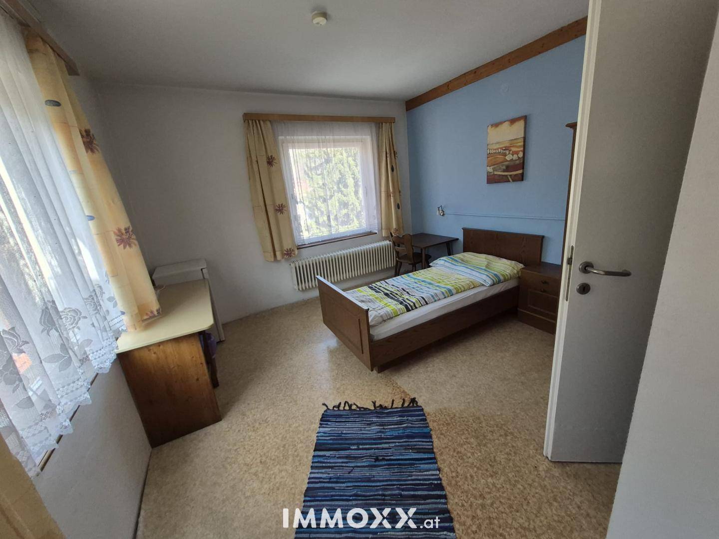 Schlafzimmer mit Einzelbett, Holzmöbeln und Fenster mit Vorhängen.