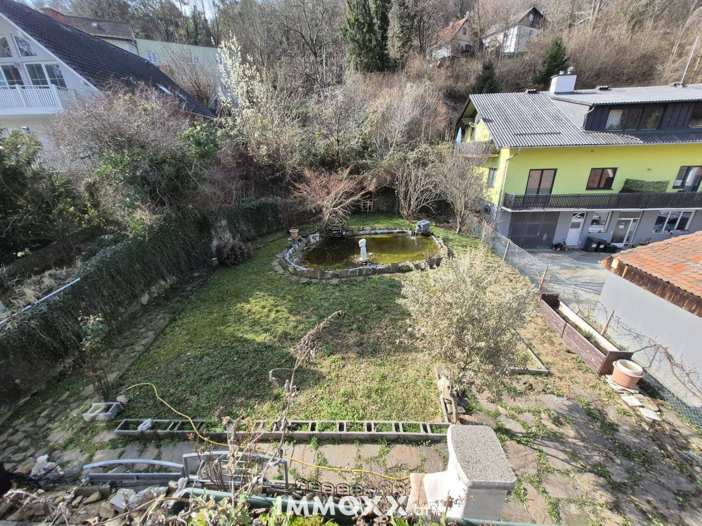 Gartenbereich mit Teich und Rasenfläche, umgeben von Bäumen und Sträuchern.