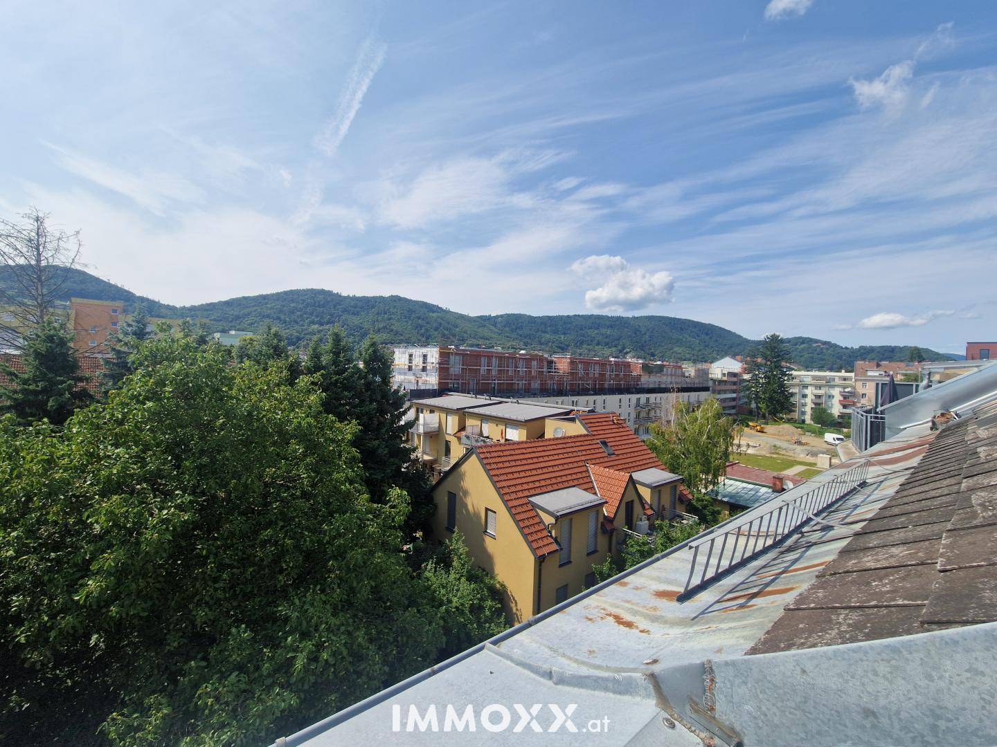 Weitläufiger Blick über Dächer, Bäume und die umliegende Hügellandschaft unter blauem Himmel.