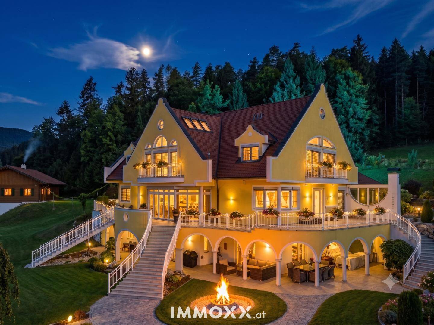 Beleuchtete Villa bei Nacht mit gemütlicher Terrasse und Feuerstelle unter dem Mondschein.