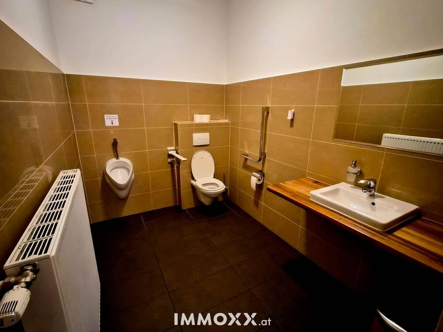 Barrierefreies WC mit Urinal, Toilette, Waschbecken und Haltegriffen an braunen Fliesenwänden.