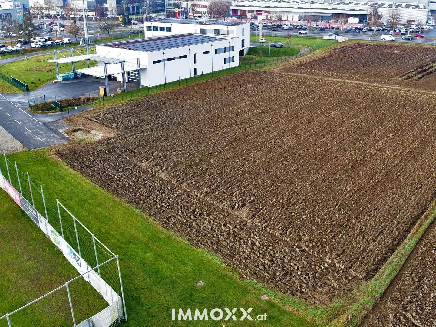 Luftaufnahme eines Ackerfeldes neben einem modernen Gebäude mit Solaranlage und einem Sportplatz.