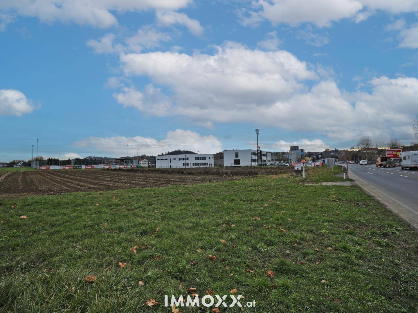 Weitläufiges Grasfeld neben einer Straße mit Gebäuden und bewölktem Himmel in der Ferne.