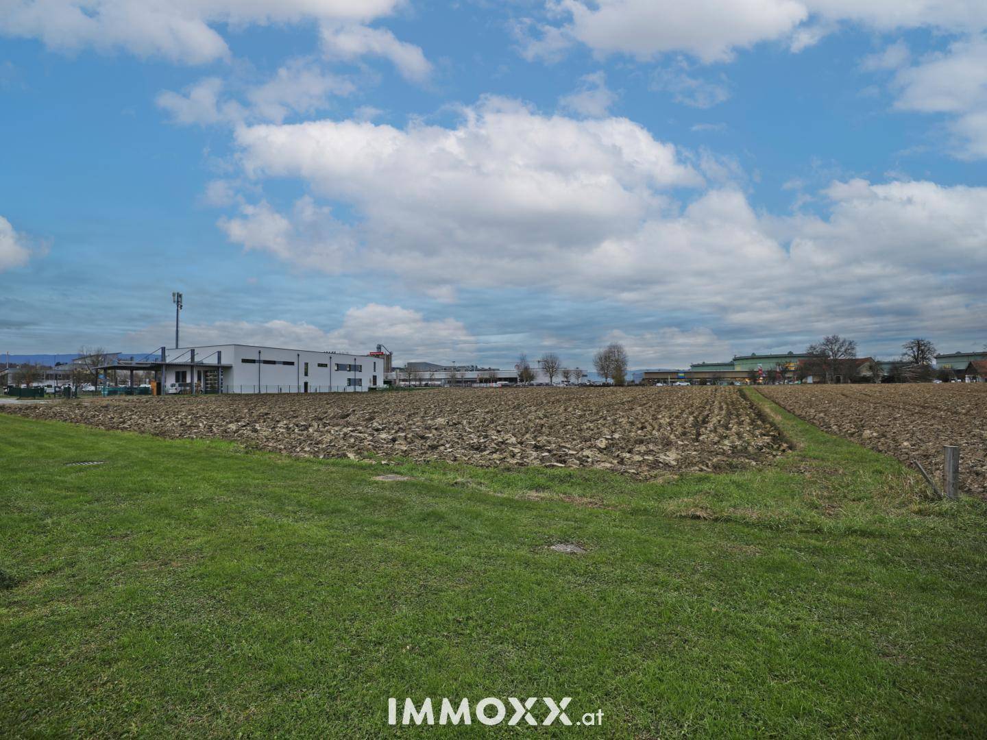 Offenes Feld mit Gras- und Ackerflächen, umgeben von entfernten Gebäuden unter einem weiten Himmel.