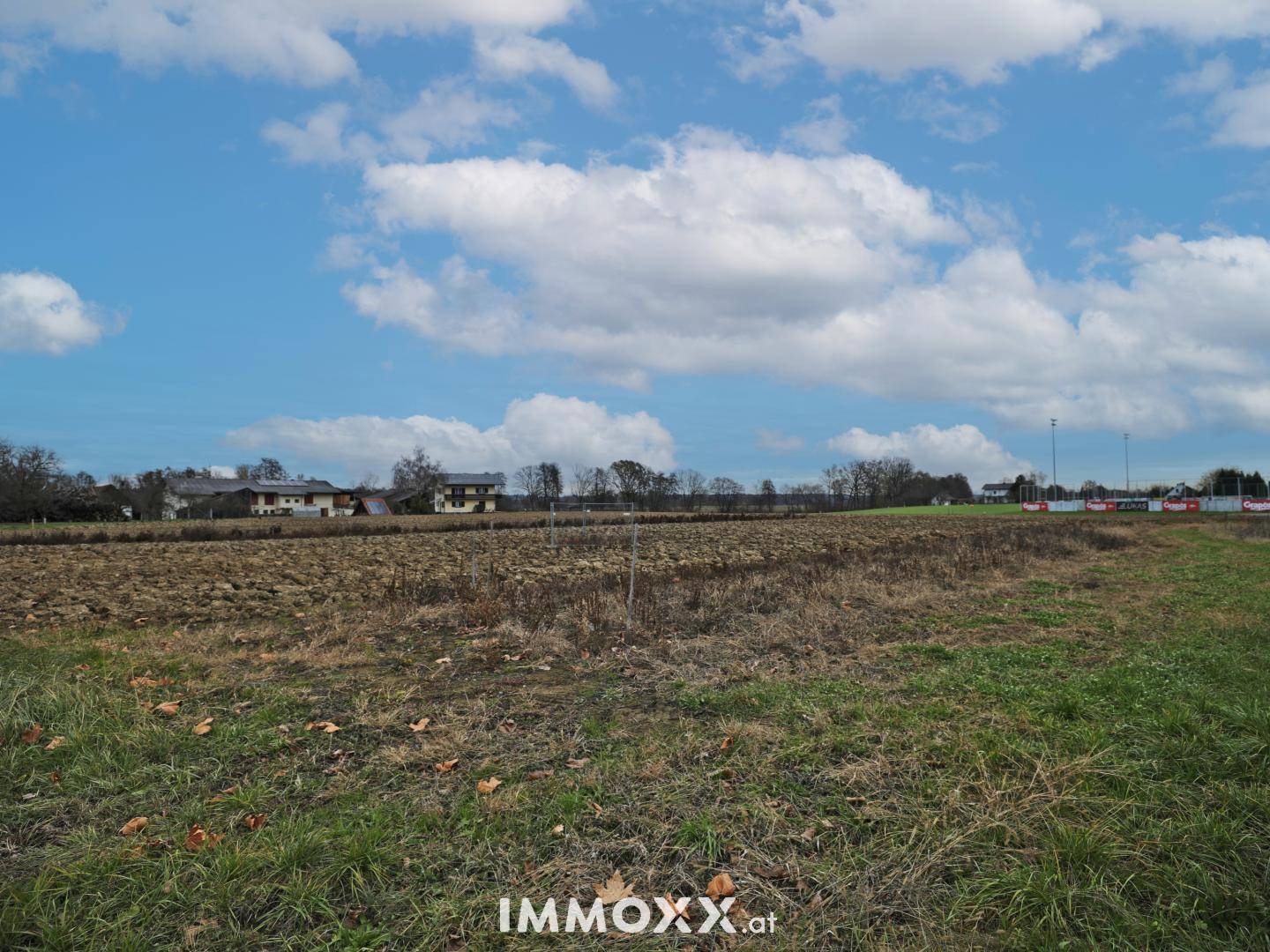 Ländliche Ansicht eines Feldes mit Gras und Ackerland, im Hintergrund sind Häuser und Bäume zu sehen.