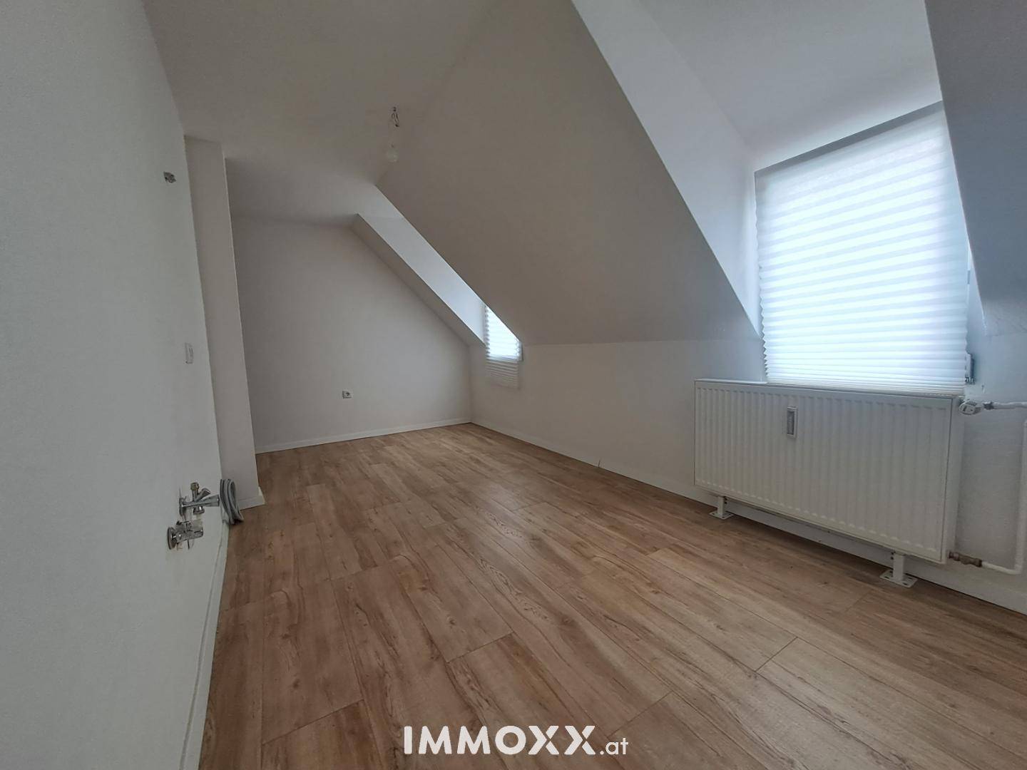 Heller Dachgeschossraum mit Laminatboden, schrägen Wänden und einem Fenster mit Jalousien.
