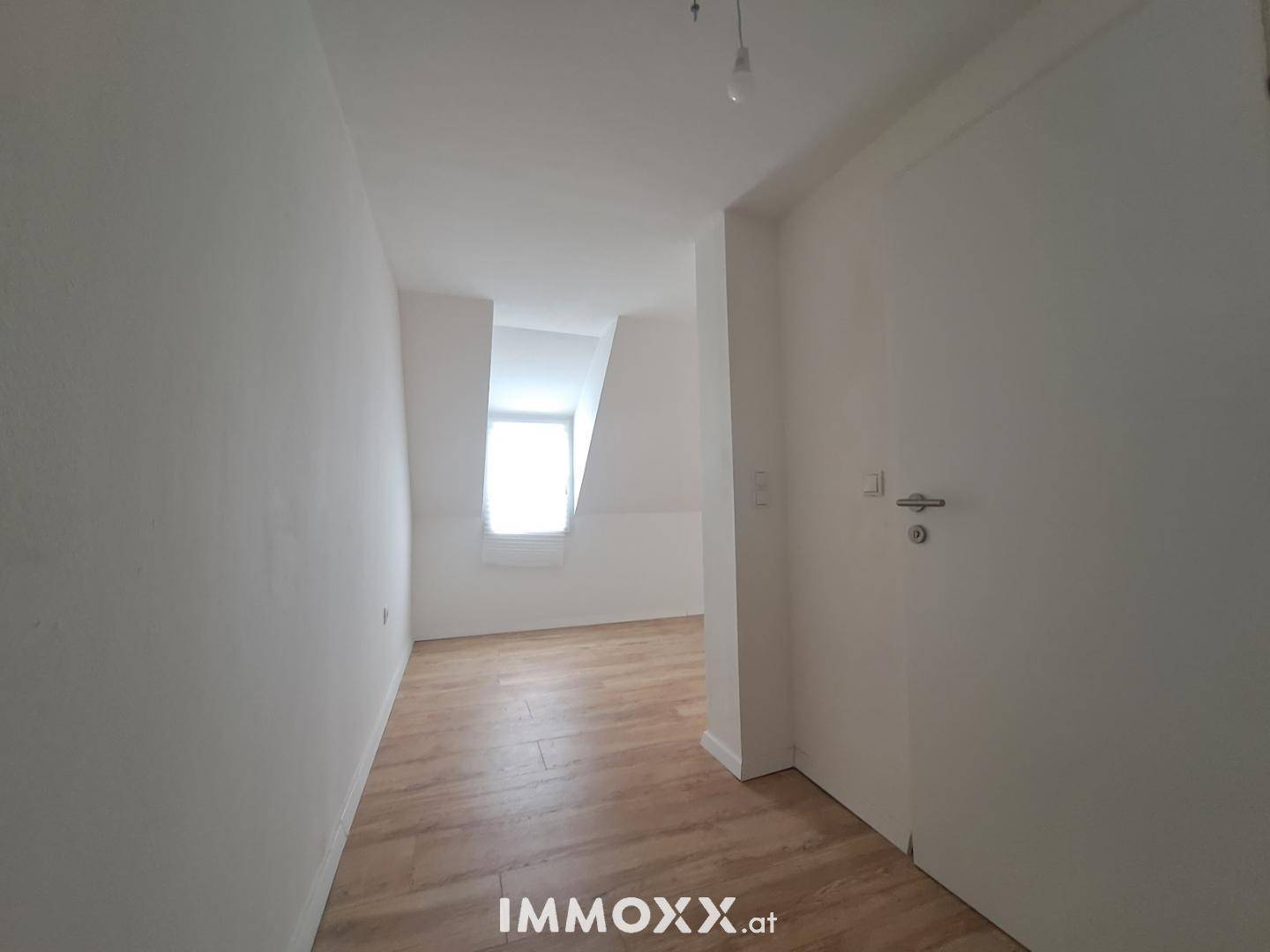 Leerer Raum im Dachgeschoss mit Laminatboden, weißen Wänden und einem Fenster in der Ferne.