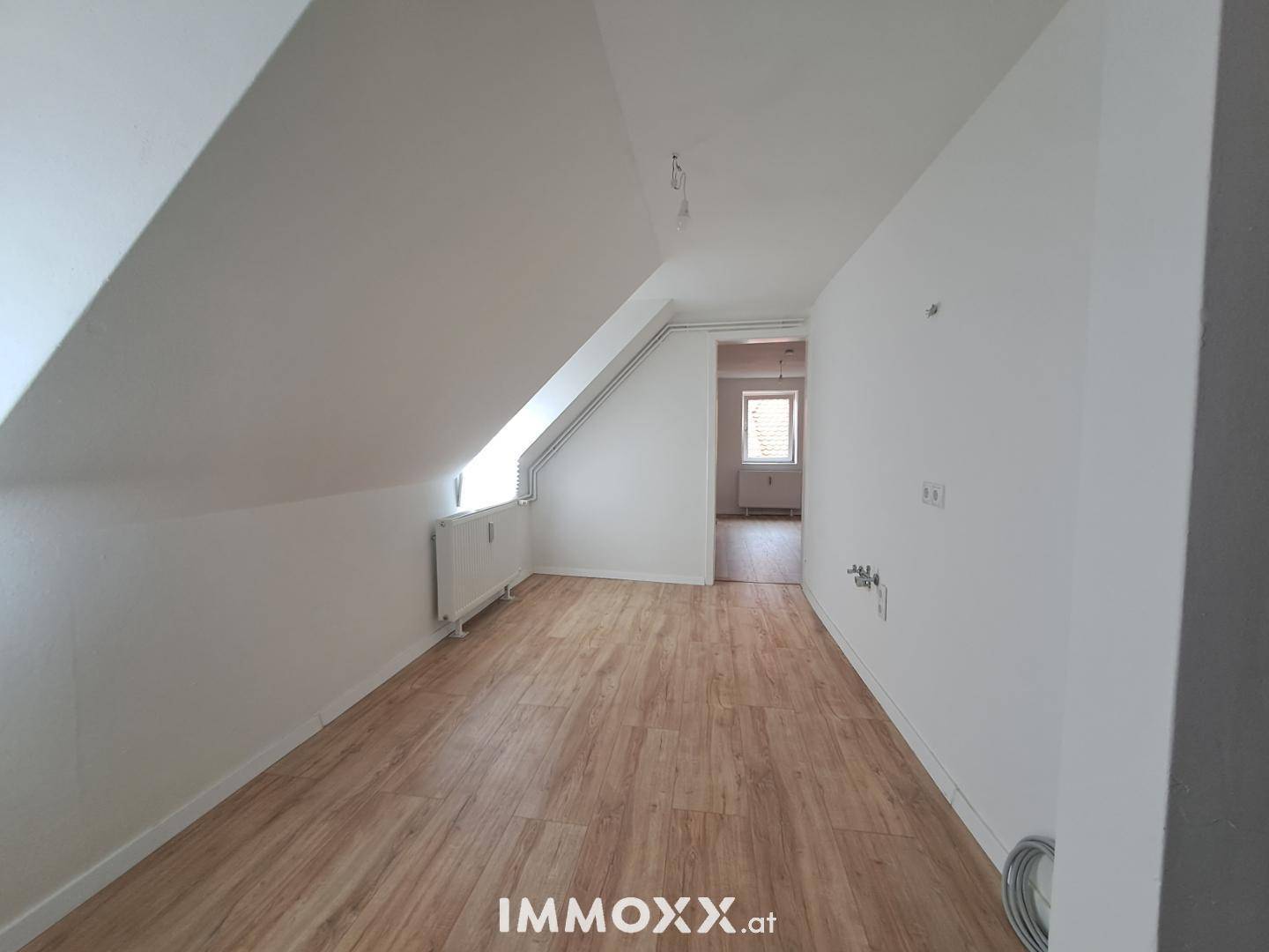 Geräumiger Dachgeschossraum mit schrägen Wänden, Laminatboden und einem Fenster mit Heizkörper.