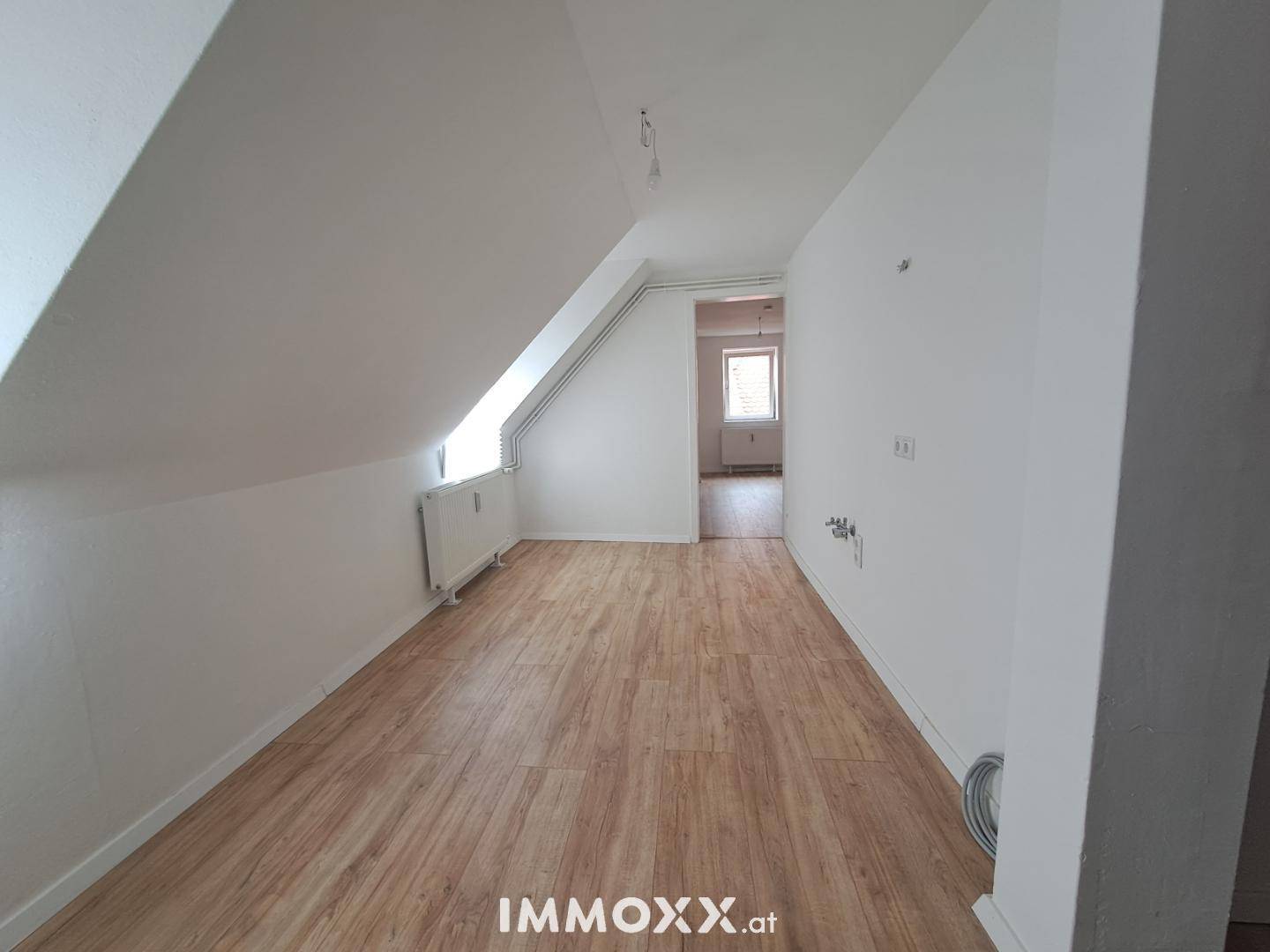 Heller Dachgeschossraum mit schrägen Wänden, Laminatboden und einem Fenster mit Heizkörper.