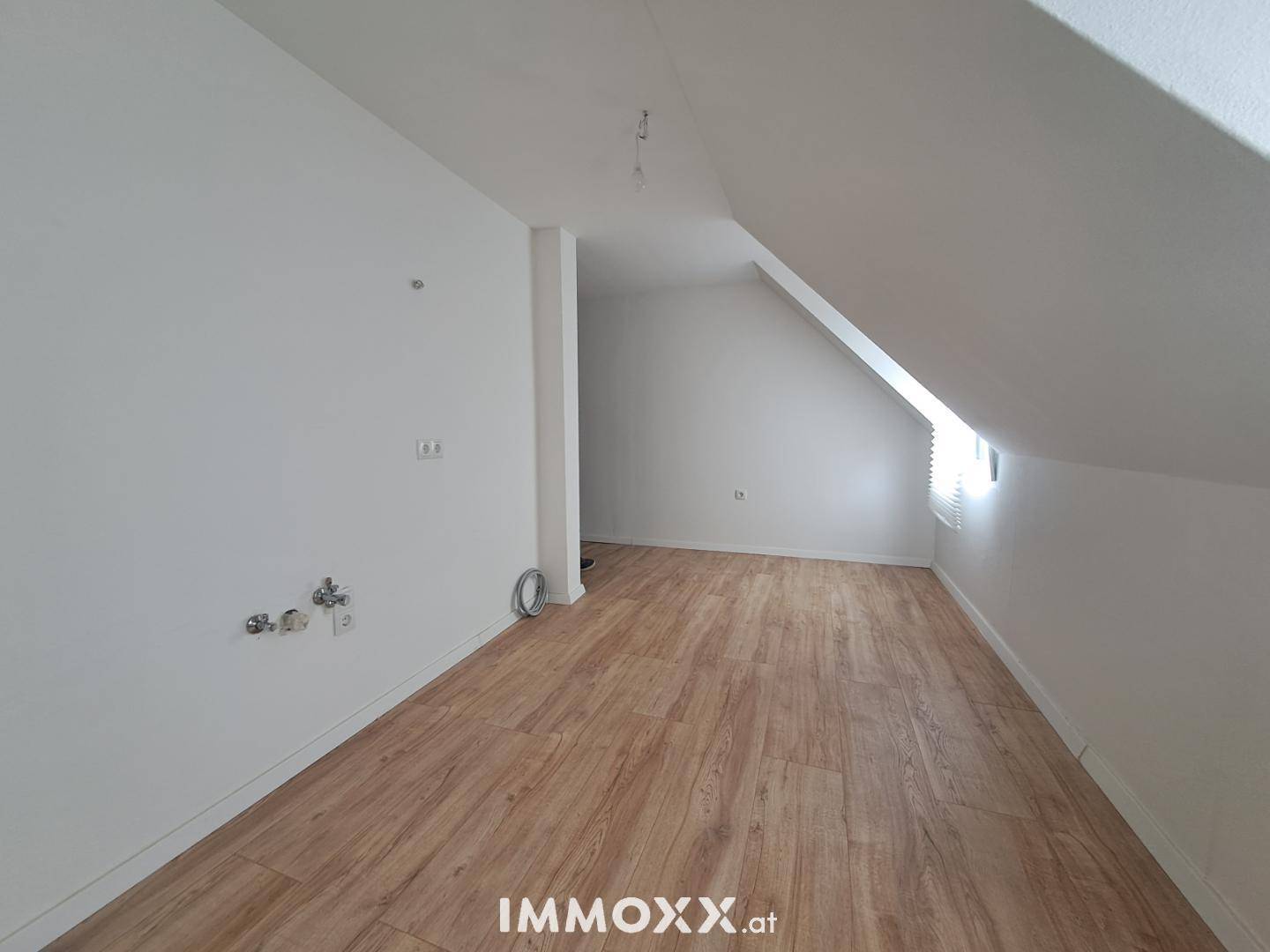 Leerer Raum im Dachgeschoss mit Laminatboden, weißen Wänden und Anschlüssen für Sanitärinstallationen.