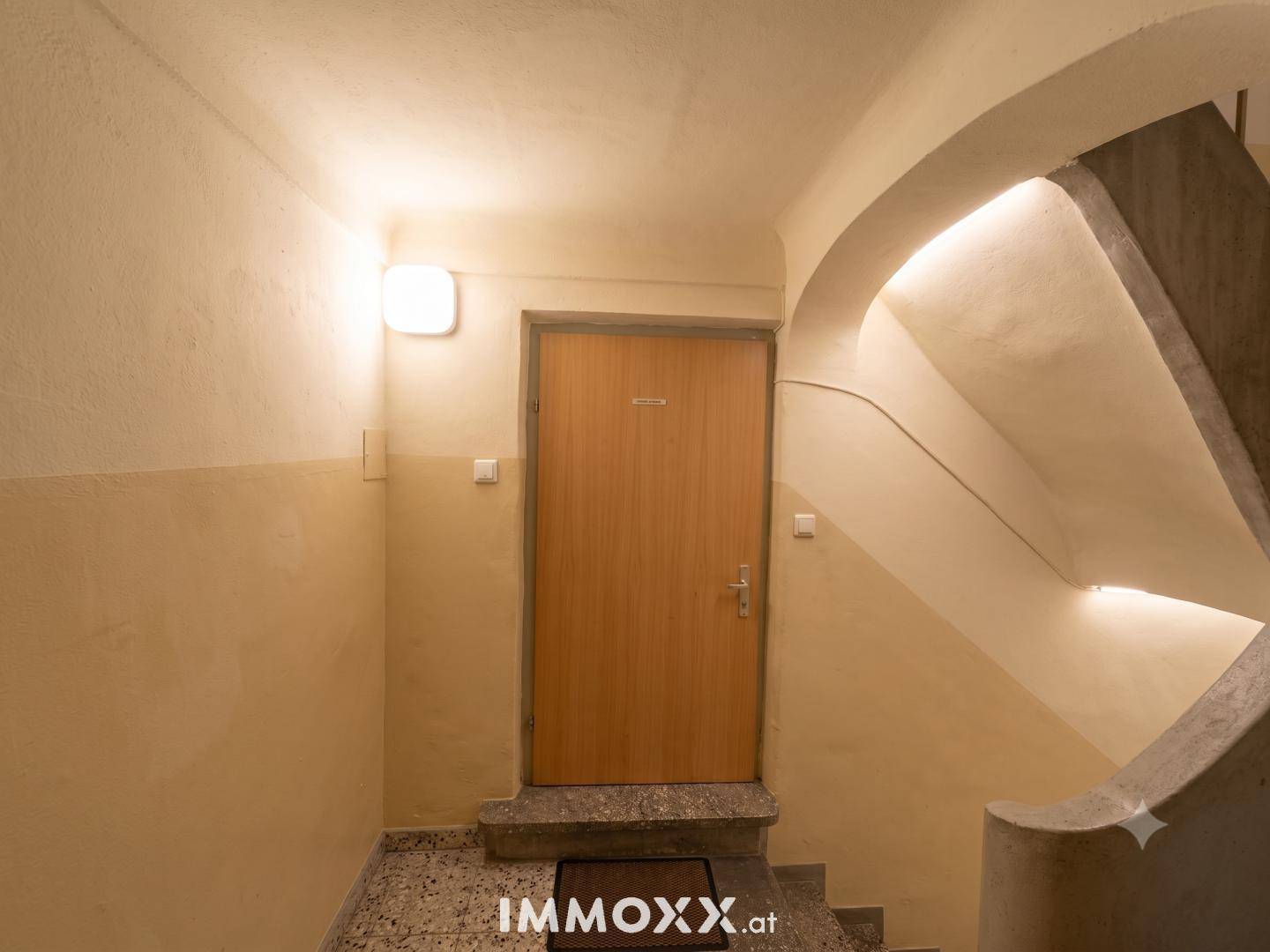 Treppenabsatz in einem Altbau mit Holztür und Terrazzoboden, der den Eingangsbereich beleuchtet.