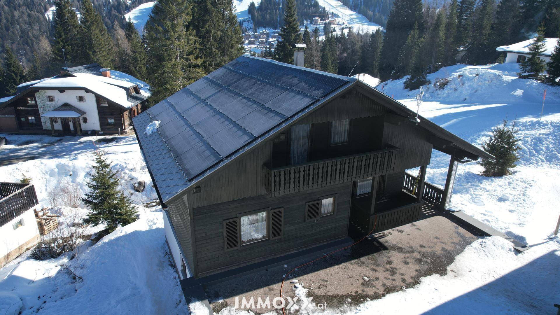 Luftaufnahme eines Holzchalets im Winter mit schneebedecktem Dach und umliegender alpiner Landschaft.