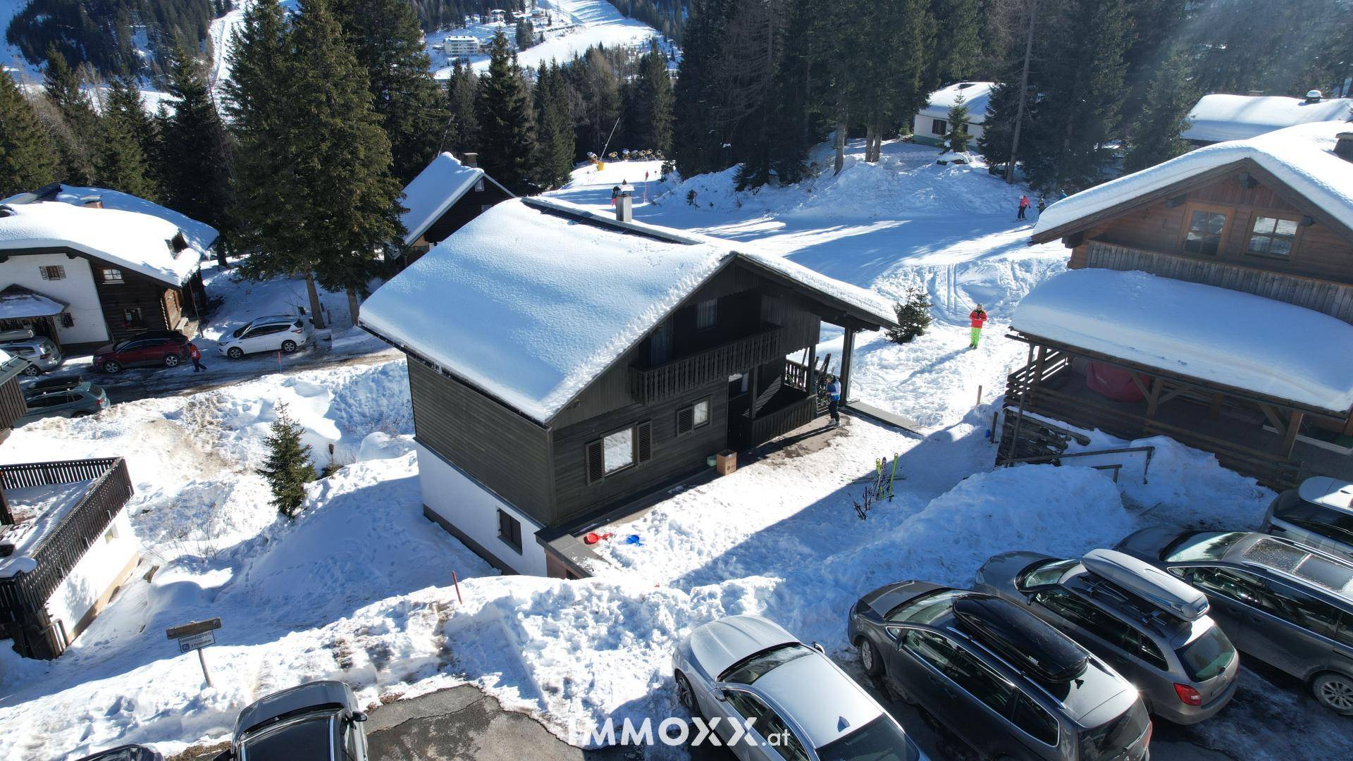 Panoramablick auf ein Holzchalet und weitere Gebäude in einer verschneiten Berglandschaft mit Parkplätzen.