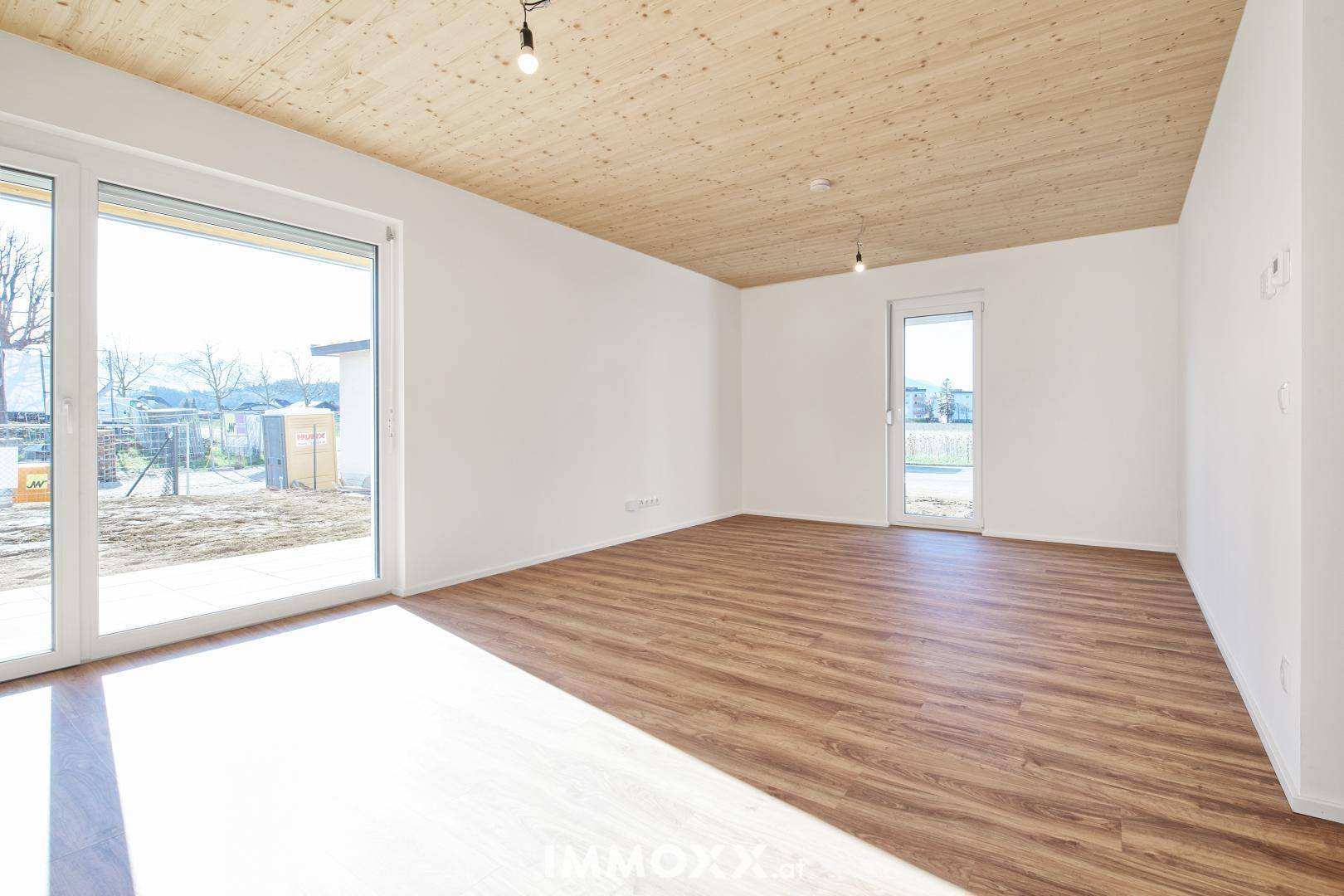 Heller, unmöblierter Raum mit Laminatboden, Holzbalkendecke und großem Fenster zum Garten.