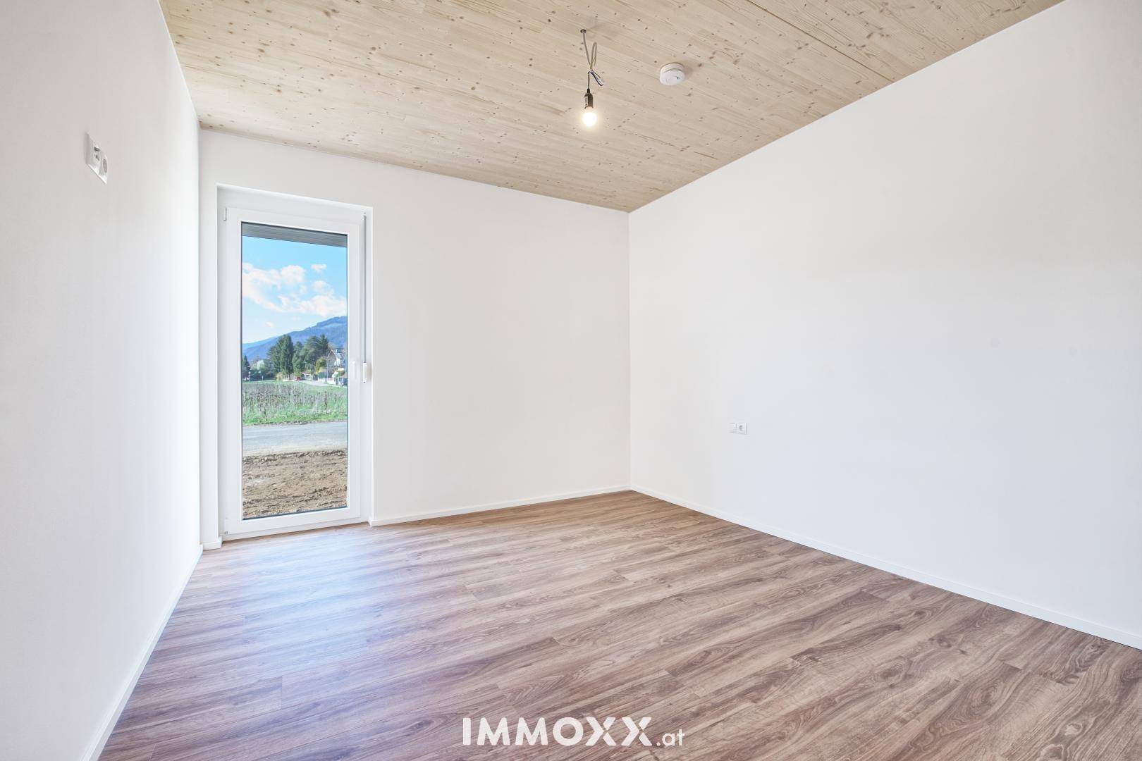 Unmöbliertes Zimmer mit Laminatboden, Holzbalkendecke und Zugang nach draußen mit Bergblick.