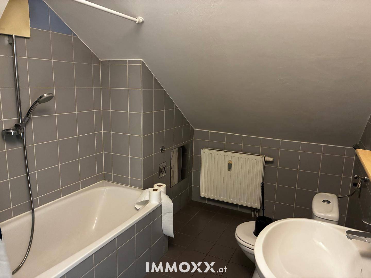 Badezimmer mit Badewanne, grauen Fliesen und schräger Decke im Dachgeschoss.
