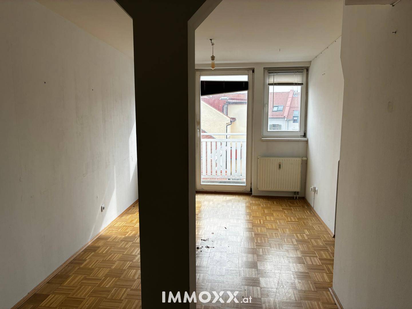 Leerer Raum mit Parkettboden, Heizkörper und Zugang zu einem kleinen Balkon.