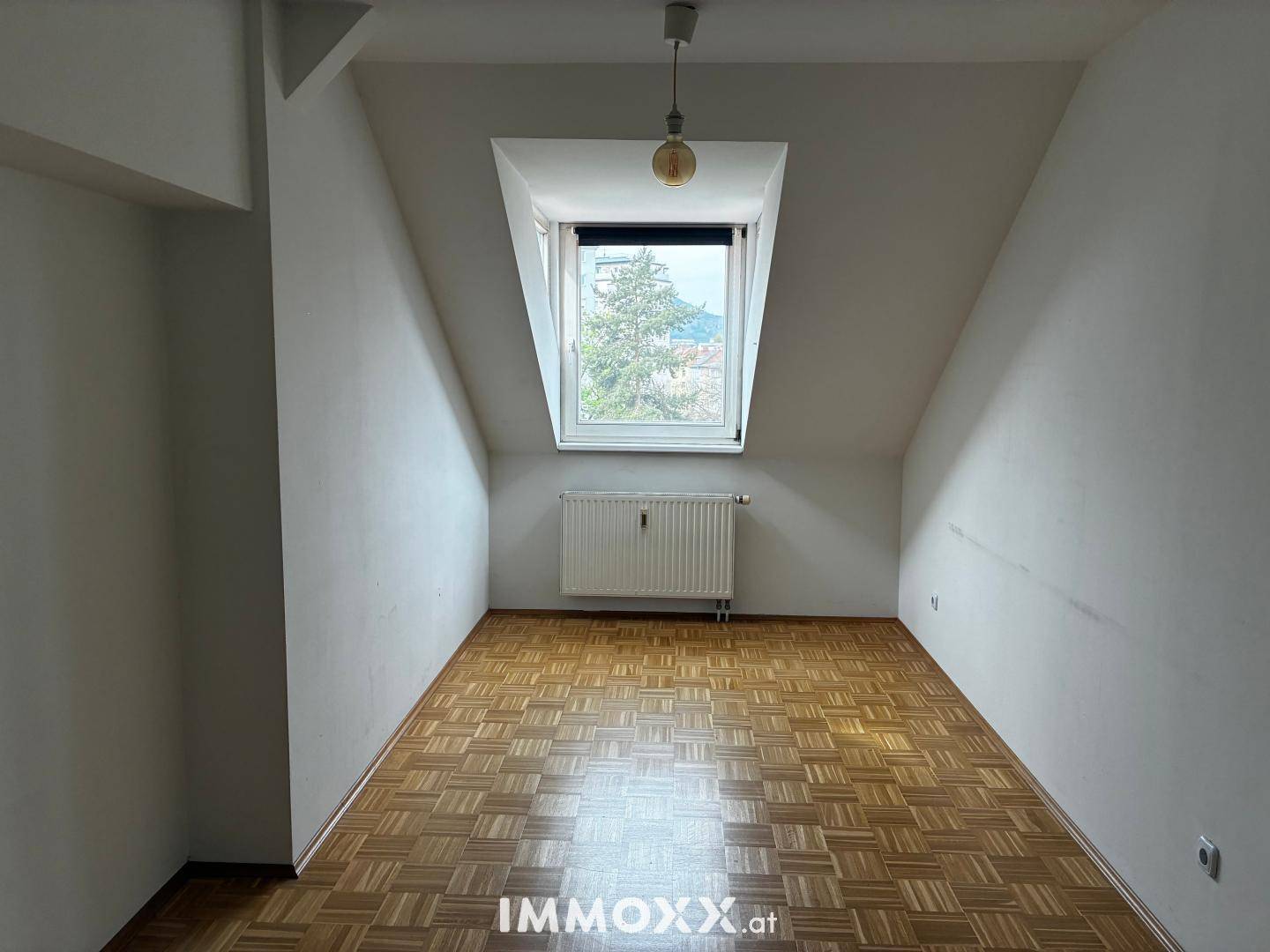 Heller Raum im Dachgeschoss mit Gaubenfenster, Heizkörper und Parkettboden.