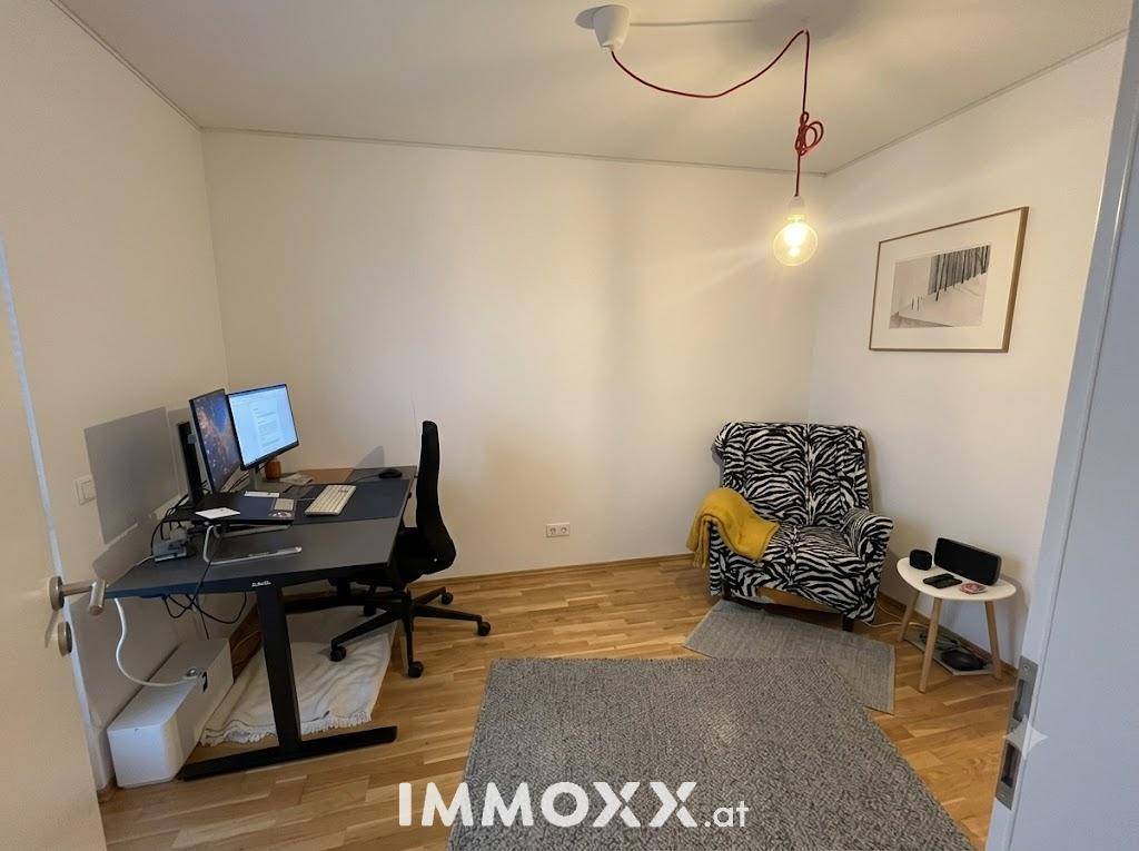 Helles Arbeitszimmer mit zwei Monitoren, Schreibtisch und einem gemusterten Sessel.