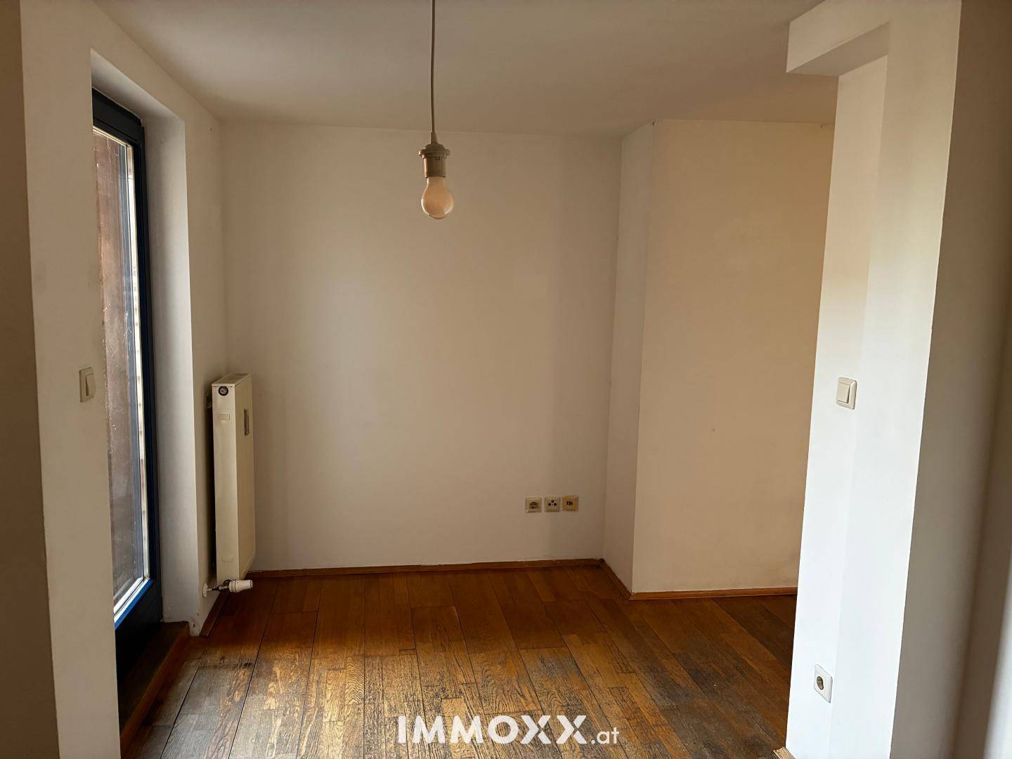 Leerer Raum mit Holzparkettboden, einem Fenster und Heizkörper, bereit zur individuellen Gestaltung.