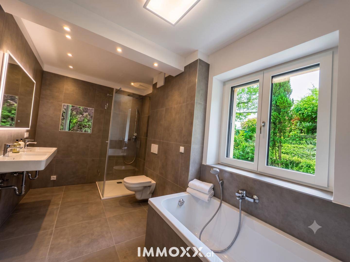 Modernes Badezimmer mit großer Badewanne, begehbarer Dusche und Fenster mit Grünblick.