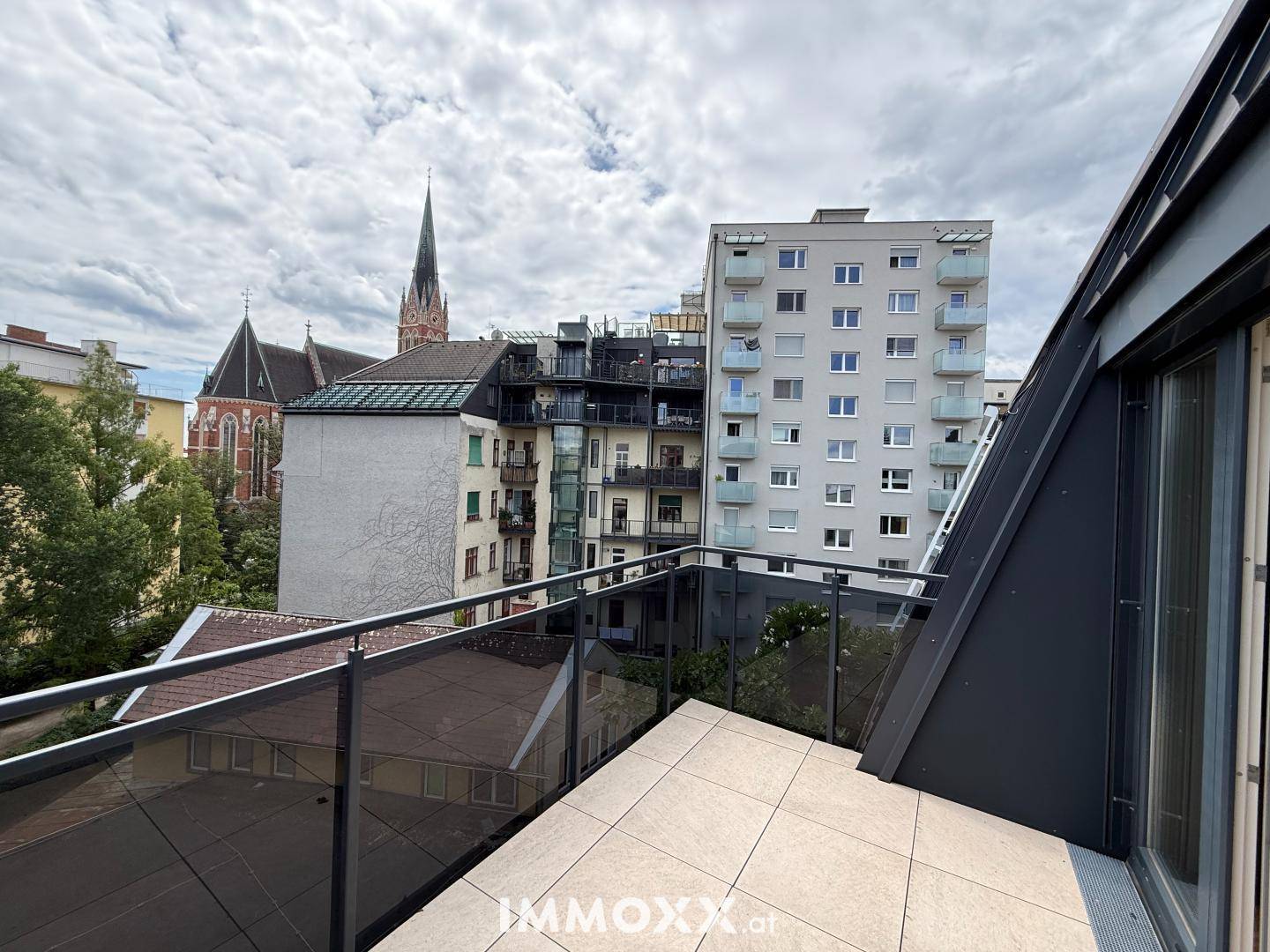 Moderne Terrasse mit Fliesenboden und Glasgeländer, bietet Blick auf die Stadt und eine Kirche.