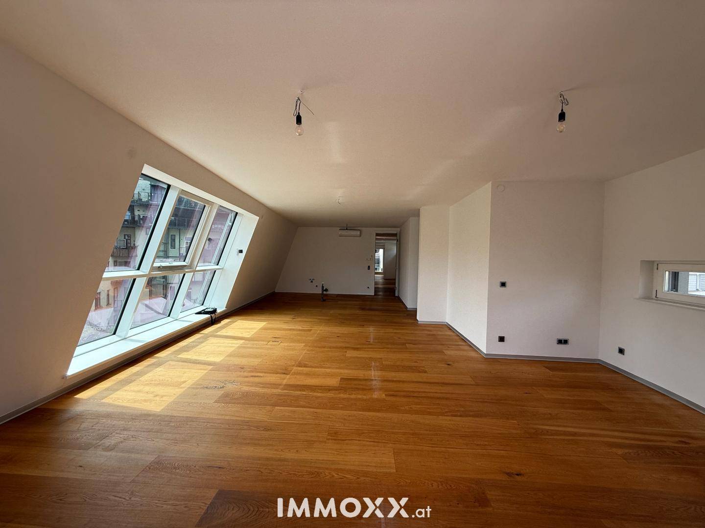 Lichtdurchfluteter, unmöblierter Raum mit elegantem Holzboden und markanten, schrägen Fensterfronten.