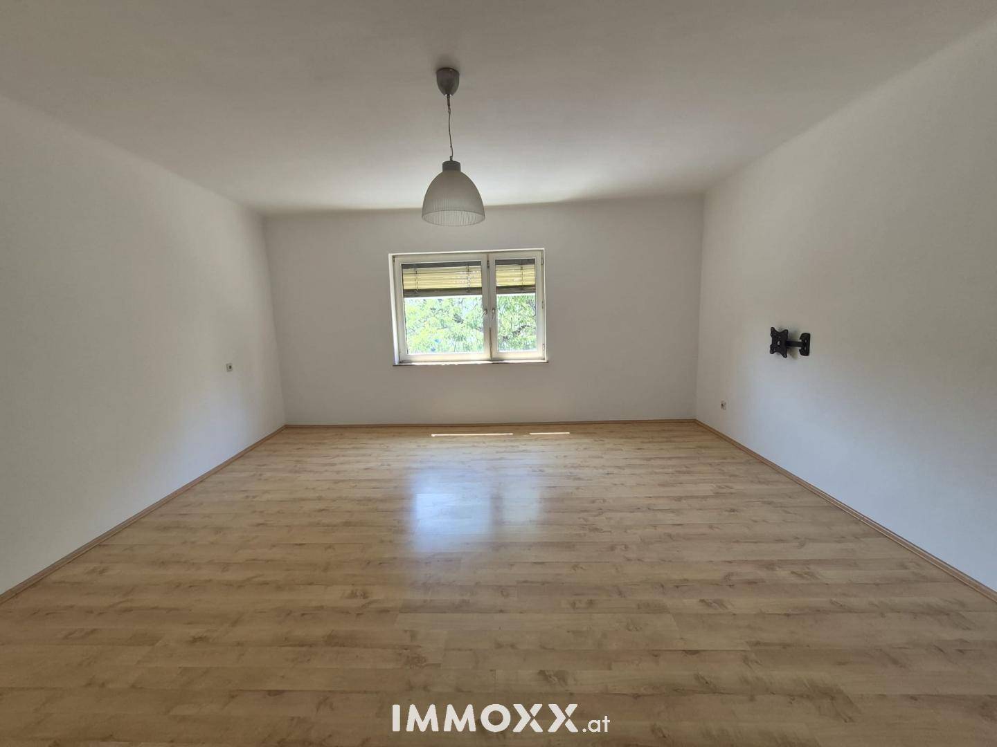 Geräumiger, leerer Raum mit Laminatboden, einem Fenster mit Jalousien und einer Deckenleuchte.