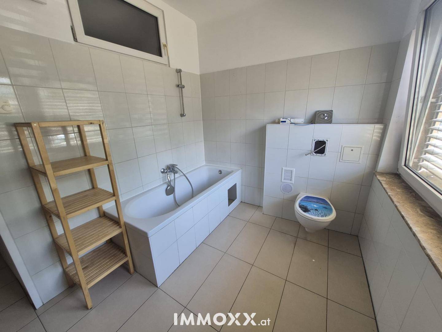 Gefliestes Badezimmer mit Badewanne und Toilette, ausgestattet mit einem Fenster für natürliche Belüftung.