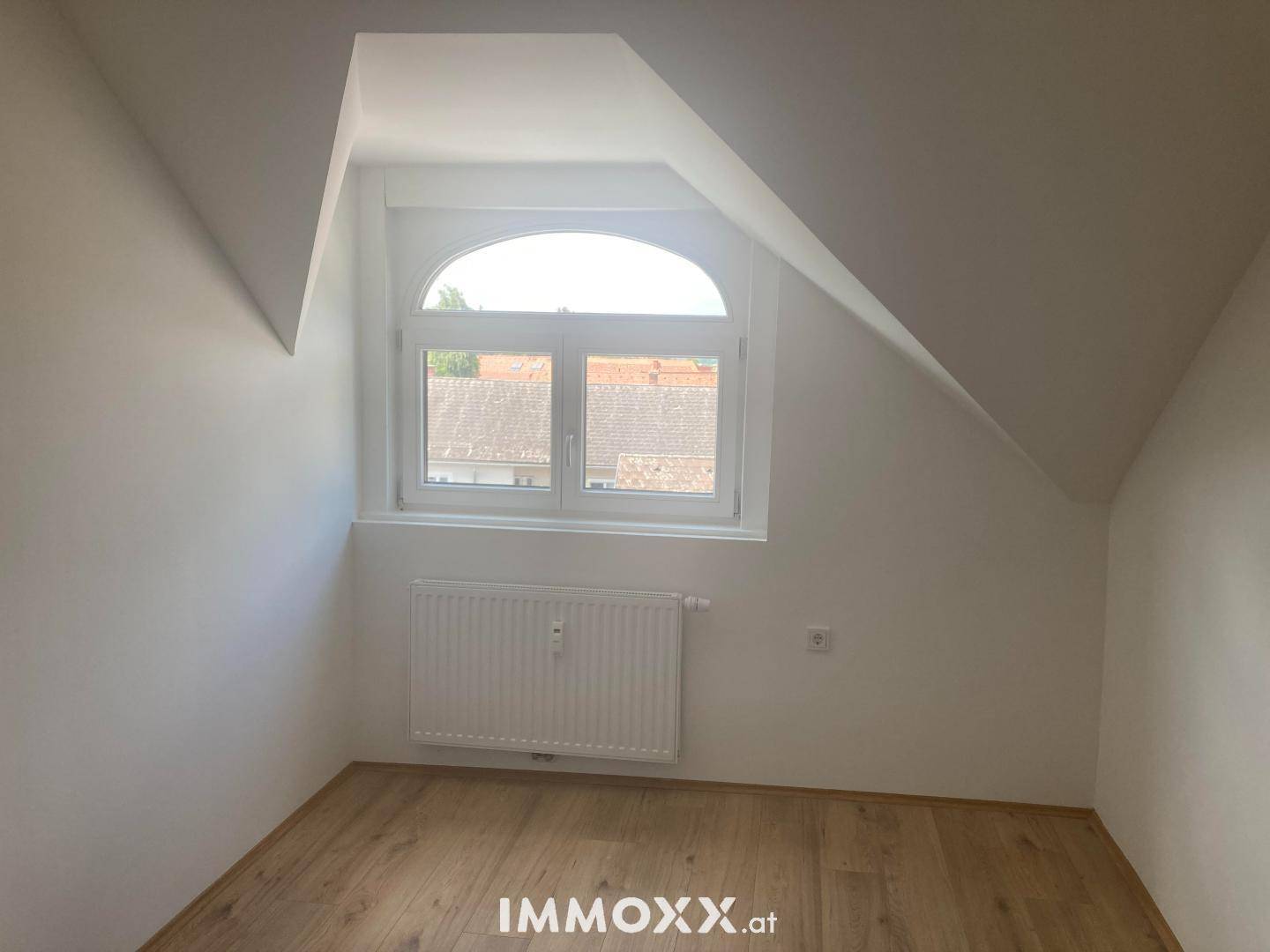 Dachgeschosszimmer mit Laminatboden, Heizkörper und einem Fenster mit Rundbogen, bietet viel Tageslicht.