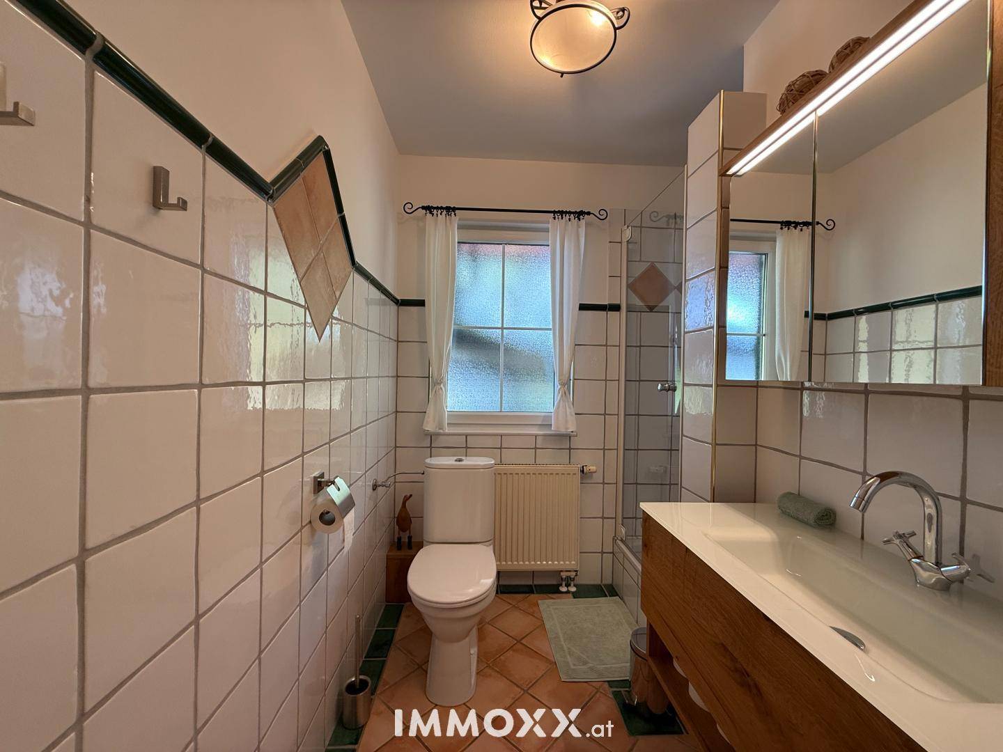 Badezimmer mit weißen Fliesen, WC, Dusche und einem Fenster für Tageslicht.