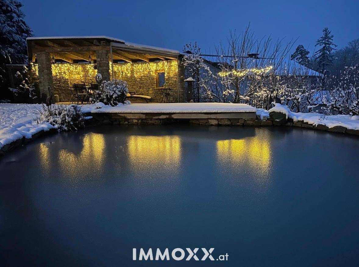 Winterliche Szene mit verschneitem Naturpool und festlich beleuchtetem Steinpavillon im Garten bei Nacht.