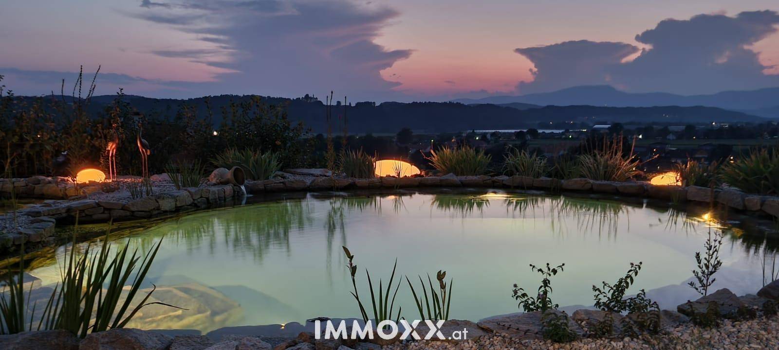Malerischer Naturpool im Garten bei Sonnenuntergang mit stimmungsvoller Beleuchtung und Bergpanorama.