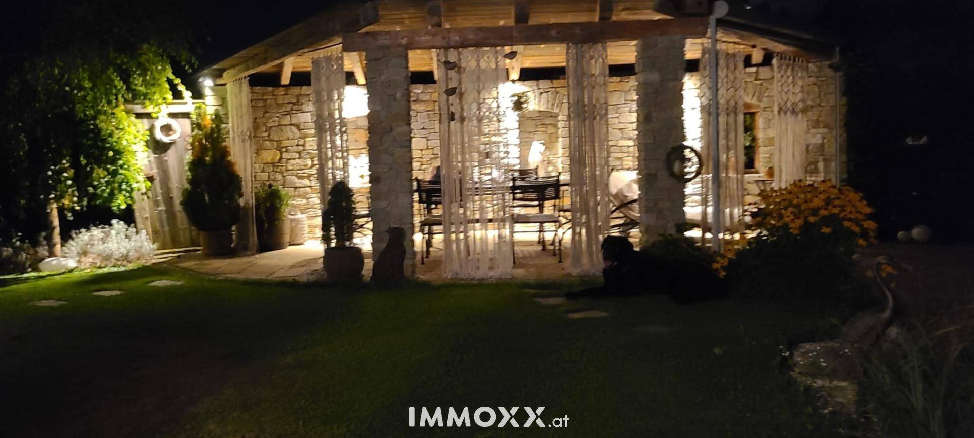 Beleuchteter Pavillon aus Naturstein im Garten mit gemütlicher Sitzecke und dekorativen Vorhängen am Abend.