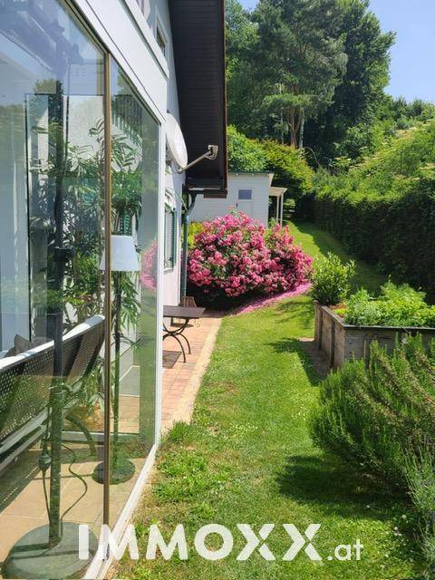 Seitlicher Blick auf das Haus mit großen Glasfronten und einem gepflegten Garten mit Blumenbeeten.