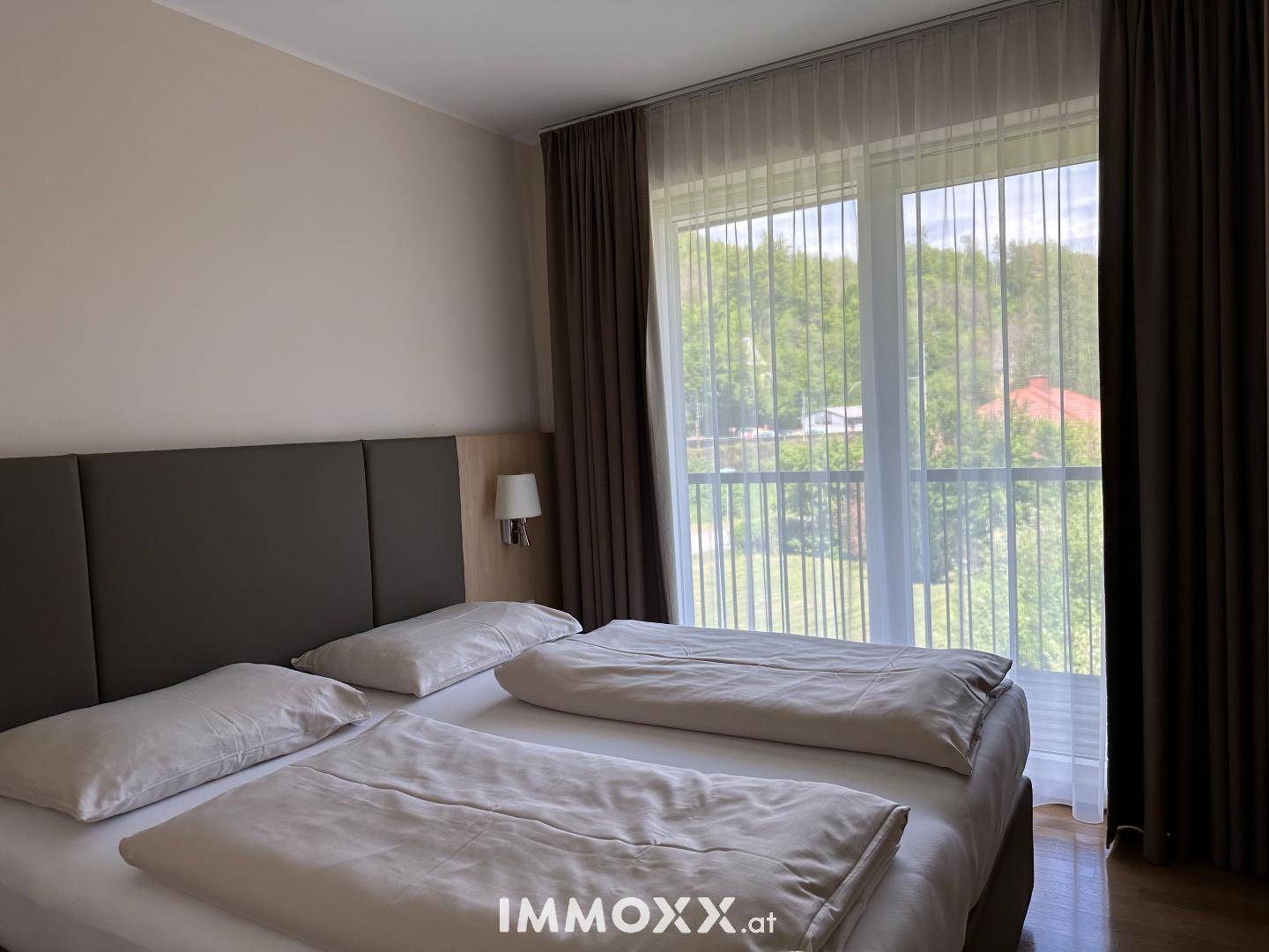 Helles Schlafzimmer mit Doppelbett und großen Fenstern, die einen grünen Ausblick bieten.