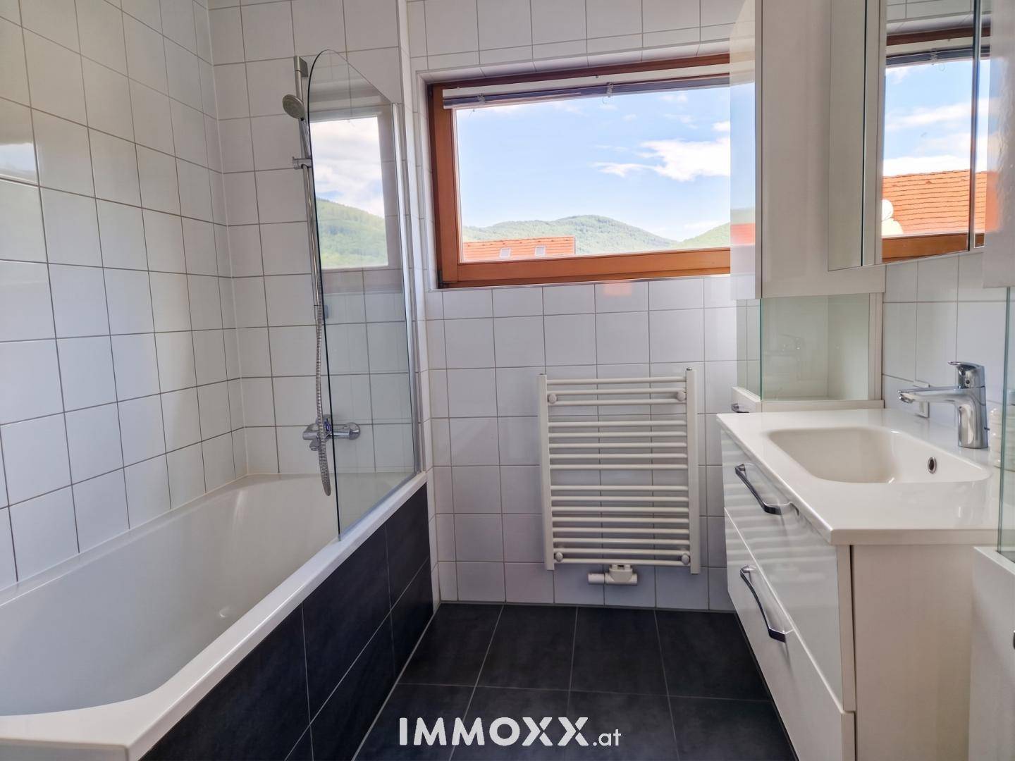 Helles Badezimmer mit Badewanne, Duschabtrennung, Waschtisch und Fenster.