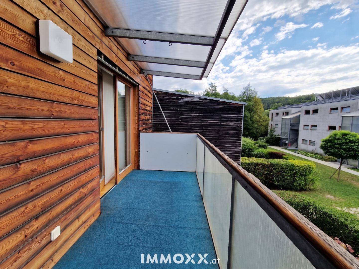 Überdachter Balkon mit blauem Teppichboden und Blick auf die grüne Umgebung.