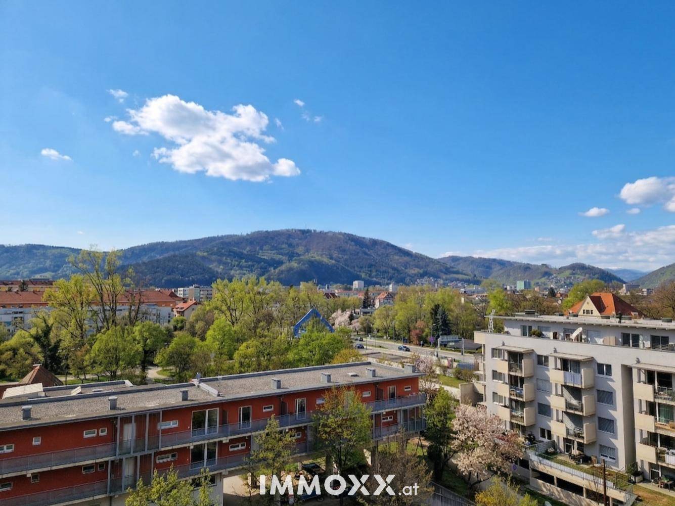 Weitblick über die Stadt und grüne Hügel mit Bergen im Hintergrund unter blauem Himmel.